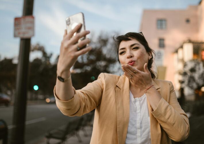Woman in a casual outfit taking a selfie on a sunny city street.