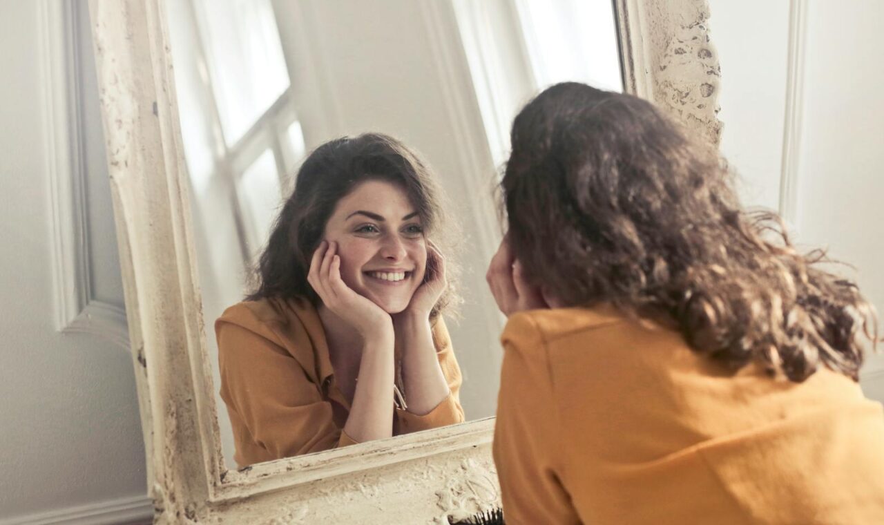 A cheerful woman smiles at her reflection in a vintage-style mirror, exuding positivity and warmth.