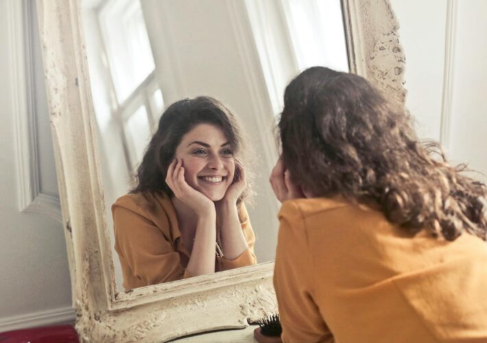 A cheerful woman smiles at her reflection in a vintage-style mirror, exuding positivity and warmth.