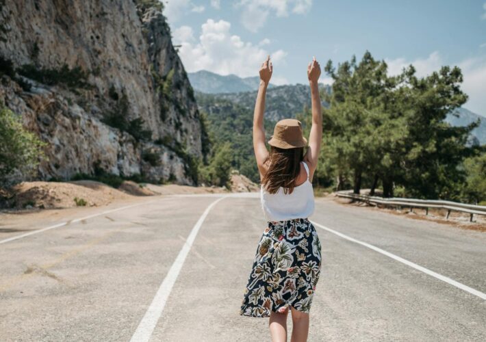 A woman with arms raised stands joyfully on a mountain road in summer.