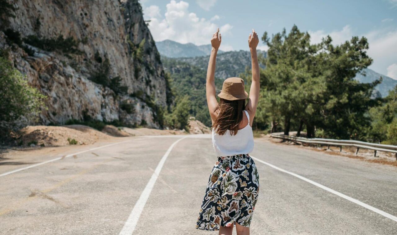 A woman with arms raised stands joyfully on a mountain road in summer.