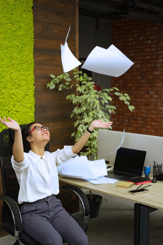 Happy Asian woman tossing papers in a modern office, celebrating success and freedom.