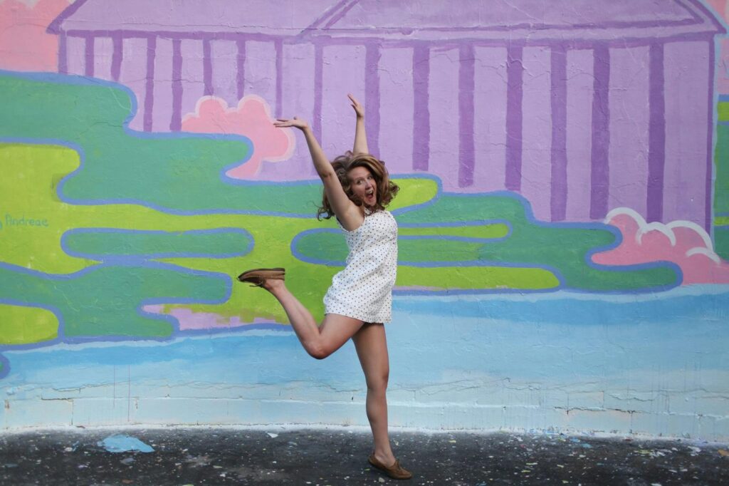 A woman joyfully jumps in front of a vibrant pastel mural, capturing a fun outdoor moment.